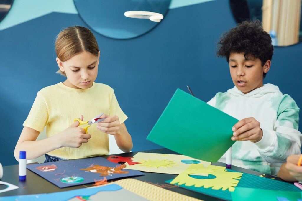 Two kids crafting together with colored papers and scissors in an art class setting.